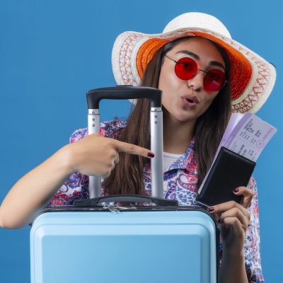 young beautiful woman tourist wearing summer hat and red sunglasses holding travel suitcase and tickets with passport pointing with finger to the looking positive and happy standing over blue b